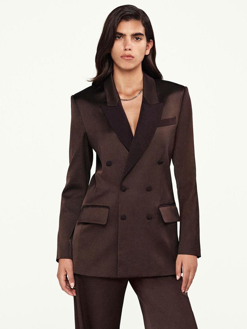 Woman in a double-breasted brown satin blazer and pants, posing against a bright backdrop in a fashion editorial shot.