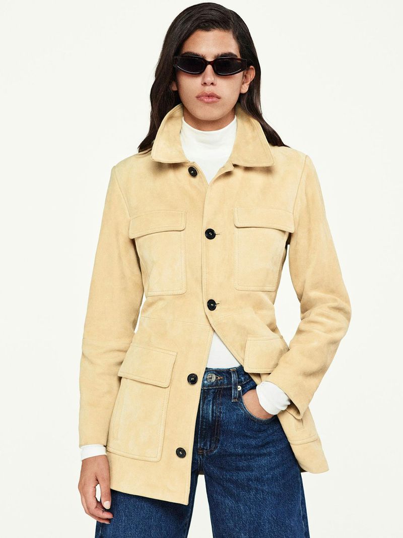 Adult female poses in a tan suede jacket, blue jeans, and dark sunglasses against a white backdrop in a studio setting.