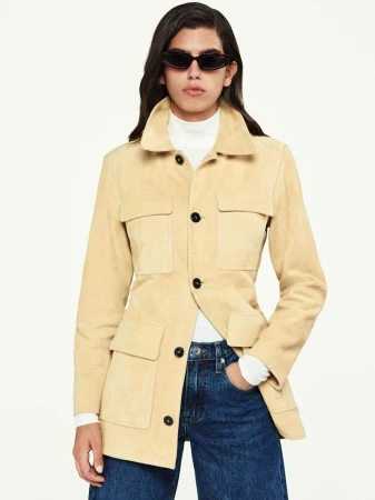 Adult female poses in a tan suede jacket, blue jeans, and dark sunglasses against a white backdrop in a studio setting.