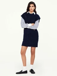 Model wearing a navy knit vest over a striped shirt and skirt with white socks and black loafers against white seamless background