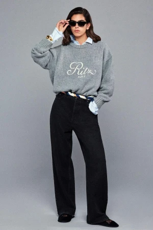 Model Sun Mizrahi wearing a grey crewneck sweater and black jeans adjustments sunglasses against a grey studio backdrop.