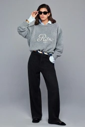 Model Sun Mizrahi wearing a grey crewneck sweater and black jeans adjustments sunglasses against a grey studio backdrop.