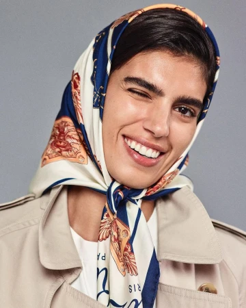 Portrait of Sun Mizrahi winking, wearing a blue and white patterned silk headscarf and a beige trench coat against a solid grey background