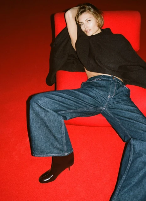 A woman in a black top and wide-leg jeans reclines on a red chair in front of a red backdrop in a fashion studio shot.