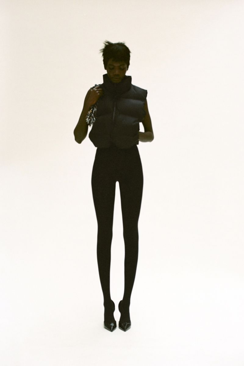A fashion shot features a slender woman with short hair wearing a black puffer vest, leggings, and heels against a bright white backdrop.
