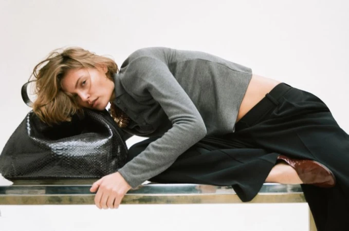 A woman reclines on a silver surface beside a large, black textured purse. She wears a grey shirt and black pants in a fashion setting.