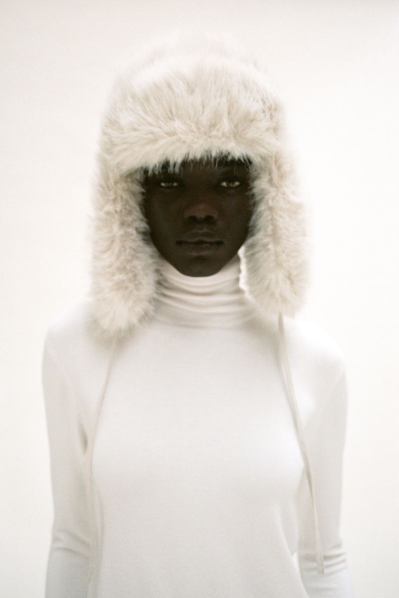 Headshot of a dark-skinned woman in a white turtleneck and a furry white hat against a bright white background in a portrait orientation.