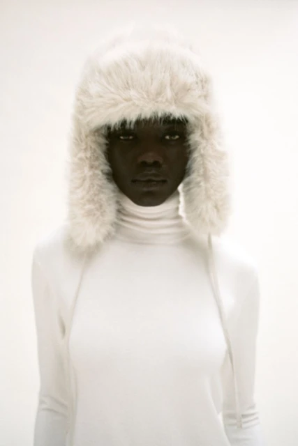 Headshot of a dark-skinned woman in a white turtleneck and a furry white hat against a bright white background in a portrait orientation.