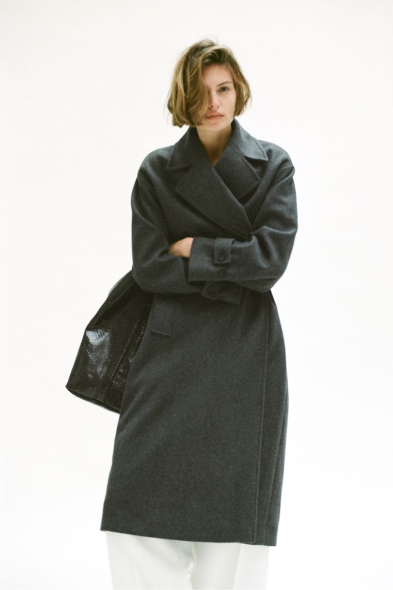 Model with short brown hair wearing a dark grey coat and white trousers standing against a white backdrop