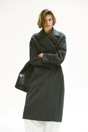 Model with short brown hair wearing a dark grey coat and white trousers standing against a white backdrop