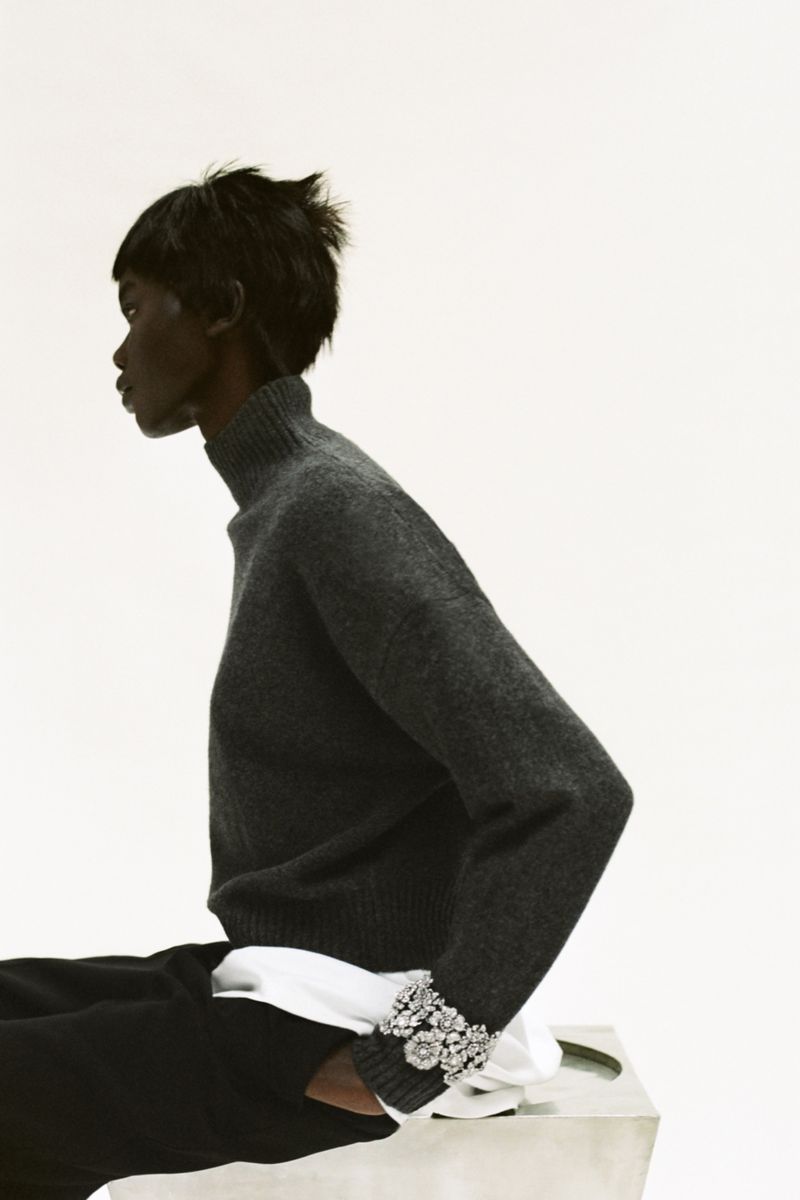 Profile of a woman with short dark hair wearing a gray turtleneck sweater and dark pants, seated against a white background.