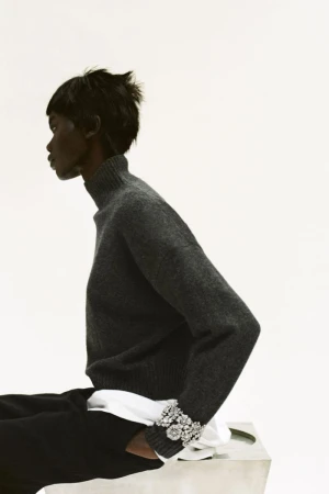 Profile of a woman with short dark hair wearing a gray turtleneck sweater and dark pants, seated against a white background.