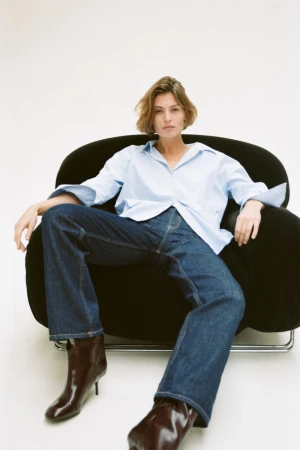 Model seated in a black armchair wearing a light blue button-down shirt and dark denim jeans against a white seamless background