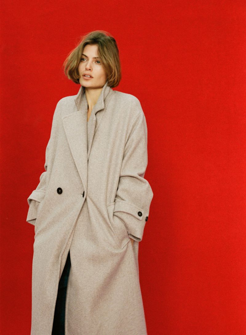 Model in a long beige belted coat posing against a solid vibrant red background.