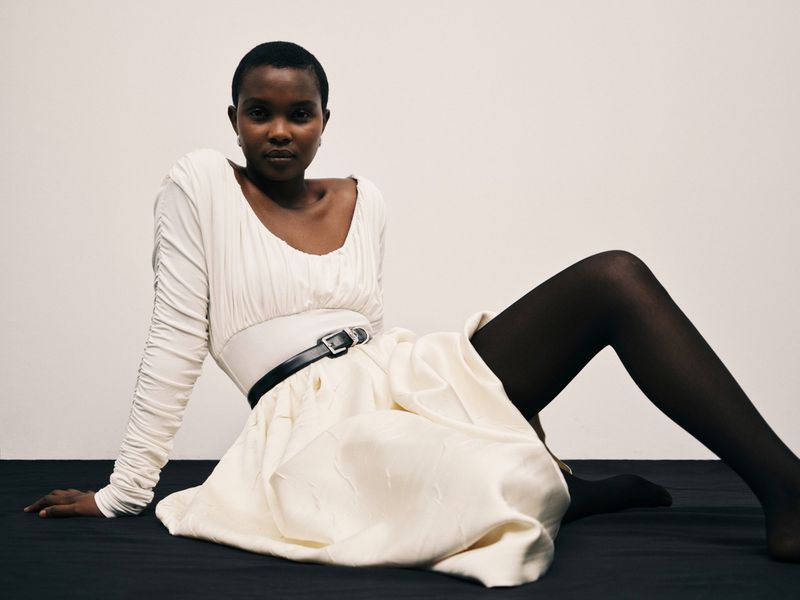 A fashionable dark-skinned model poses indoors against a white backdrop, wearing a long-sleeved dress, sheer tights, and a thin black belt.