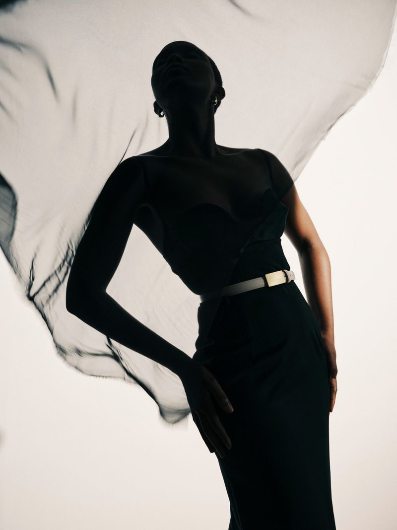 Studio shot of a silhouetted Black woman in a black dress and flowing chiffon scarf, posed against a stark white backdrop with moody lighting.
