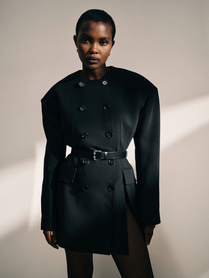 Portrait of a Black woman in a black blazer dress with a black belt, posing in front of a neutral wall with shadow.