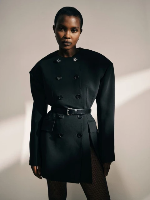 Portrait of a Black woman in a black blazer dress with a black belt, posing in front of a neutral wall with shadow.