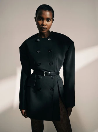 Portrait of a Black woman in a black blazer dress with a black belt, posing in front of a neutral wall with shadow.