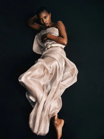 Portrait of a slender African-American woman elegantly draped in a shimmering gown lying on a dark, smooth surface in a studio setting.