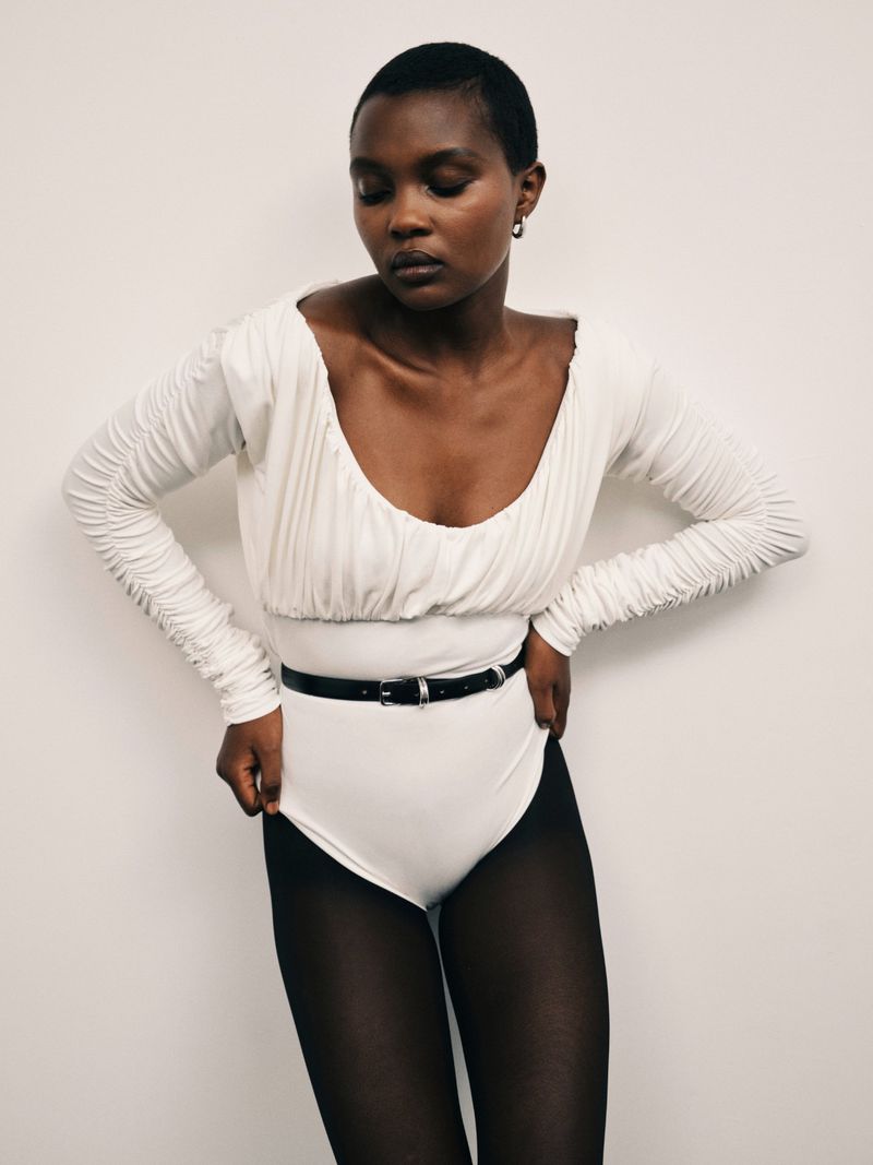 Model with short dark hair wearing a white scoop-neck top, black belt, and dark tights against a plain white background