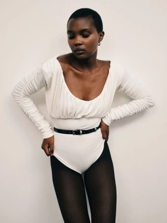 Model with short dark hair wearing a white scoop-neck top, black belt, and dark tights against a plain white background