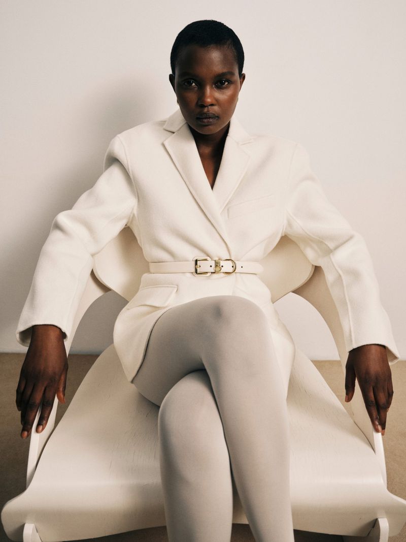 Model seated in a white chair wearing a white belted blazer and white tights against a plain background