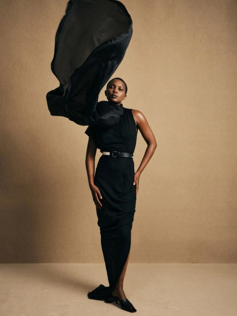 Elegant fashion shot: African woman in black dress poses gracefully with flowing fabric on neutral backdrop, showcasing style and sophistication.