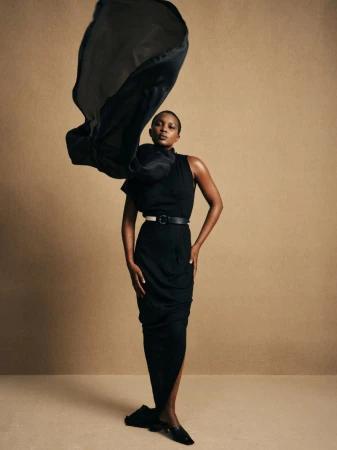 Elegant fashion shot: African woman in black dress poses gracefully with flowing fabric on neutral backdrop, showcasing style and sophistication.