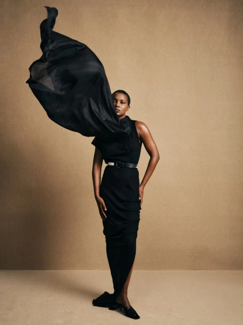 A fashionable Black woman poses in a black dress, with a dramatic black fabric swirl adding movement against a neutral backdrop in a studio shot.