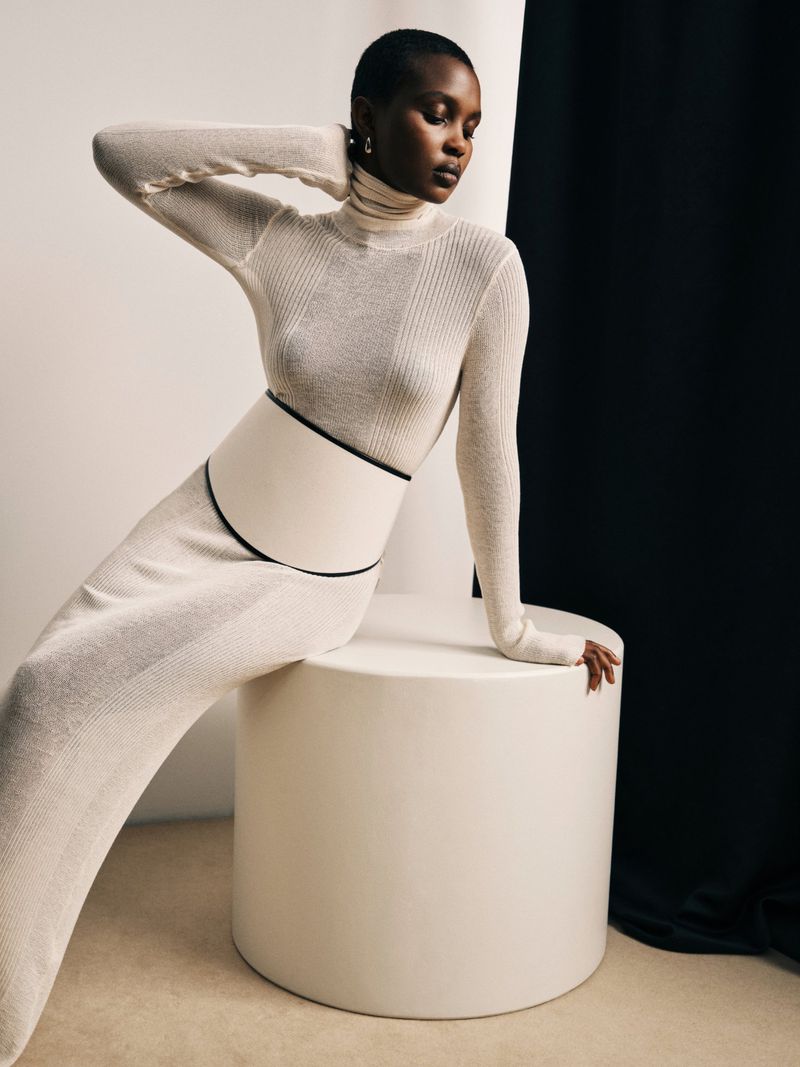 An African-American woman in a long-sleeved top and skirt poses seated on a white cylinder against a black-and-white backdrop in studio lighting.