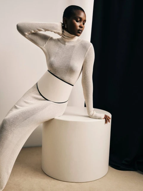 An African-American woman in a long-sleeved top and skirt poses seated on a white cylinder against a black-and-white backdrop in studio lighting.