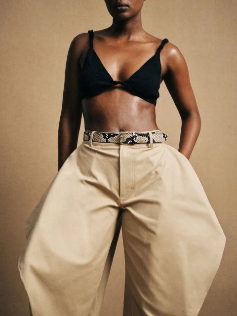 A woman with dark skin wears a black bra top and khaki wide-leg pants with a snakeskin belt in a studio shot on a beige background.