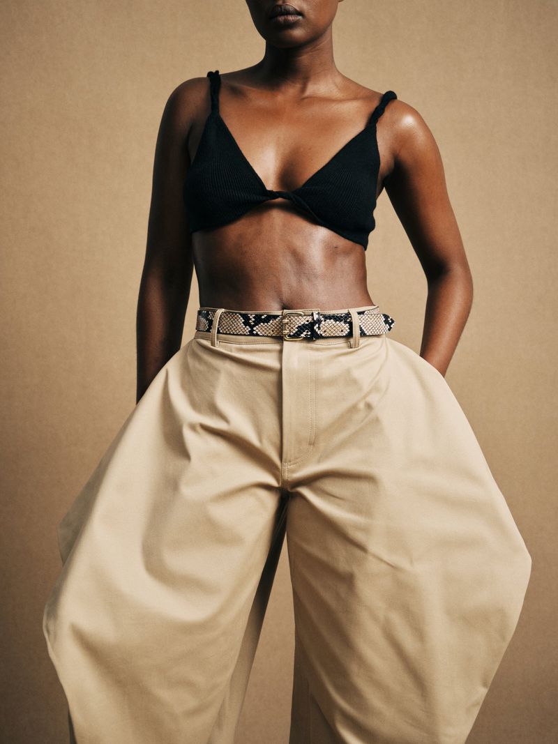 Woman in a black top and wide-legged tan pants with a snakeskin belt, standing against a neutral background in a studio shot.