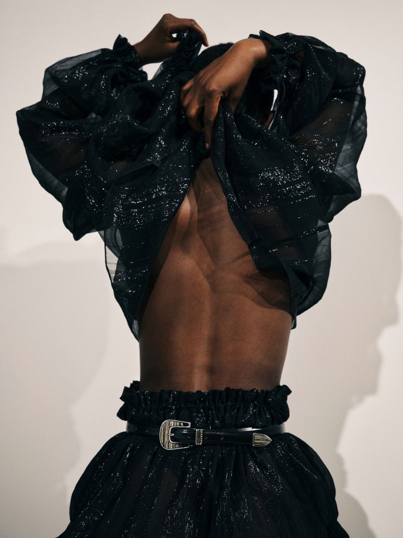 Model in a shimmering black ensemble, featuring a ruffle-trimmed top and skirt with a decorative belt and buckle, set against a plain background.