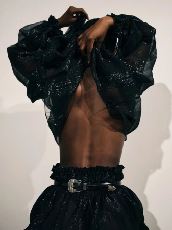 Model in a shimmering black ensemble, featuring a ruffle-trimmed top and skirt with a decorative belt and buckle, set against a plain background.
