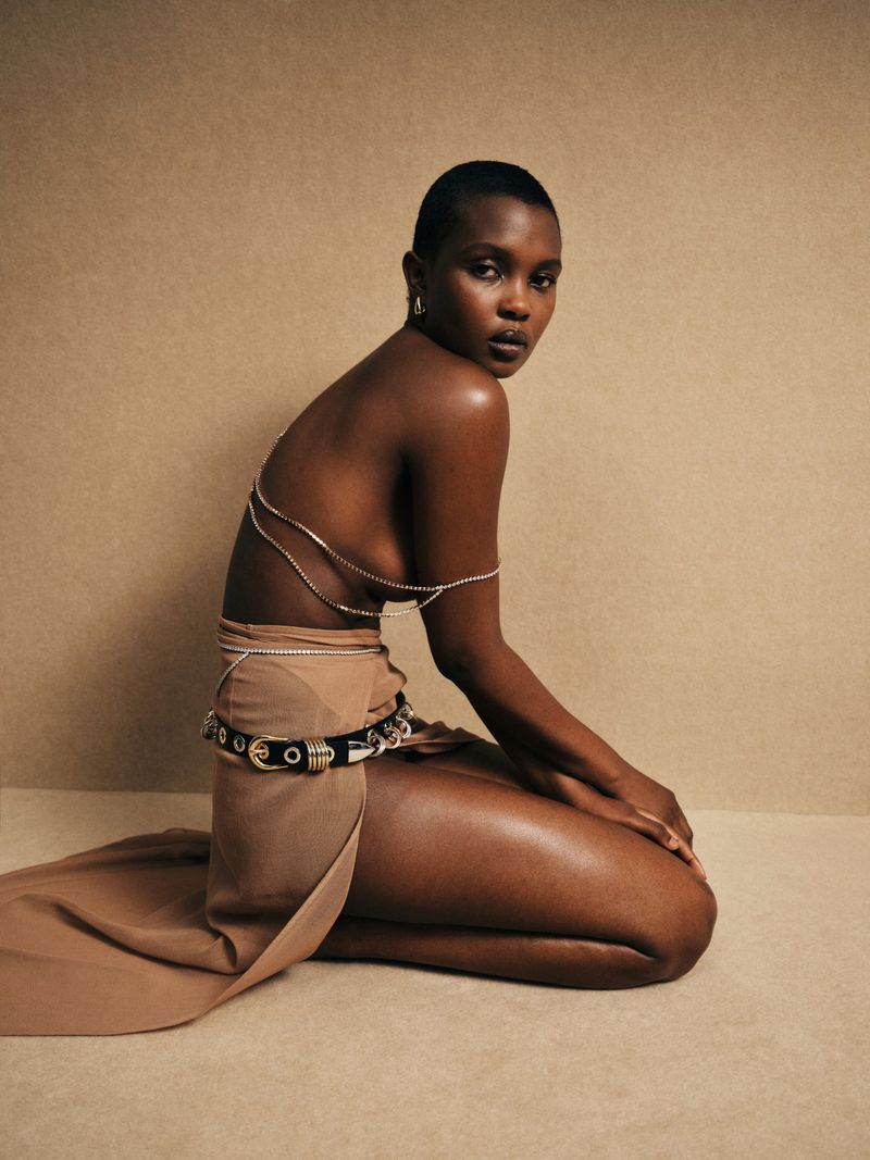 Woman with short dark hair kneeling in a tan dress with silver body jewelry and belt, against a neutral cream-colored background.