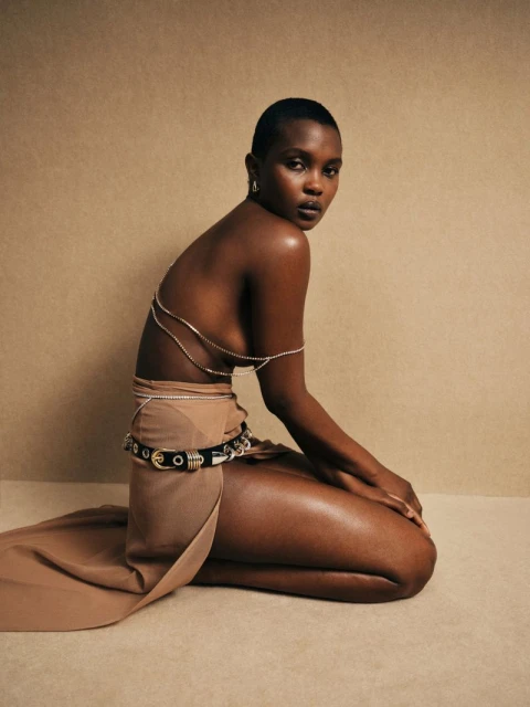 Woman with short dark hair kneeling in a tan dress with silver body jewelry and belt, against a neutral cream-colored background.