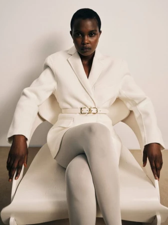Model seated in a white chair wearing a white belted blazer and white tights against a plain background