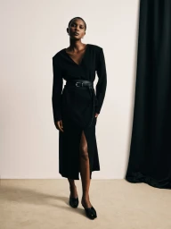 Model wearing a black belted dress and black shoes standing against a neutral studio backdrop with dark fabric curtain.