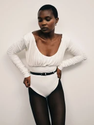 Model with short dark hair wearing a white scoop-neck top, black belt, and dark tights against a plain white background