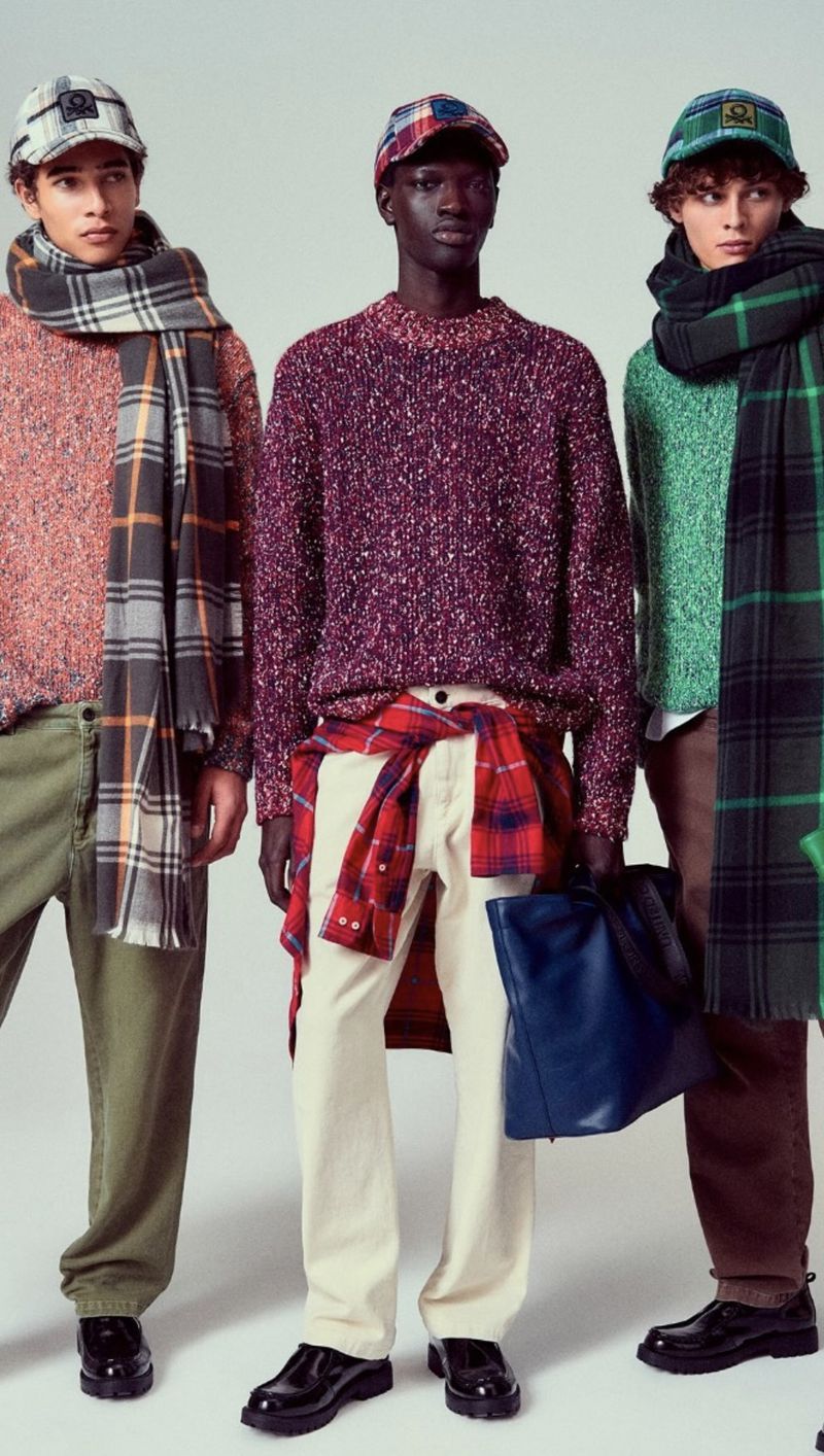 Three fashionable young adult men wearing sweaters, hats, and scarves pose indoors against a neutral background in a studio shot.