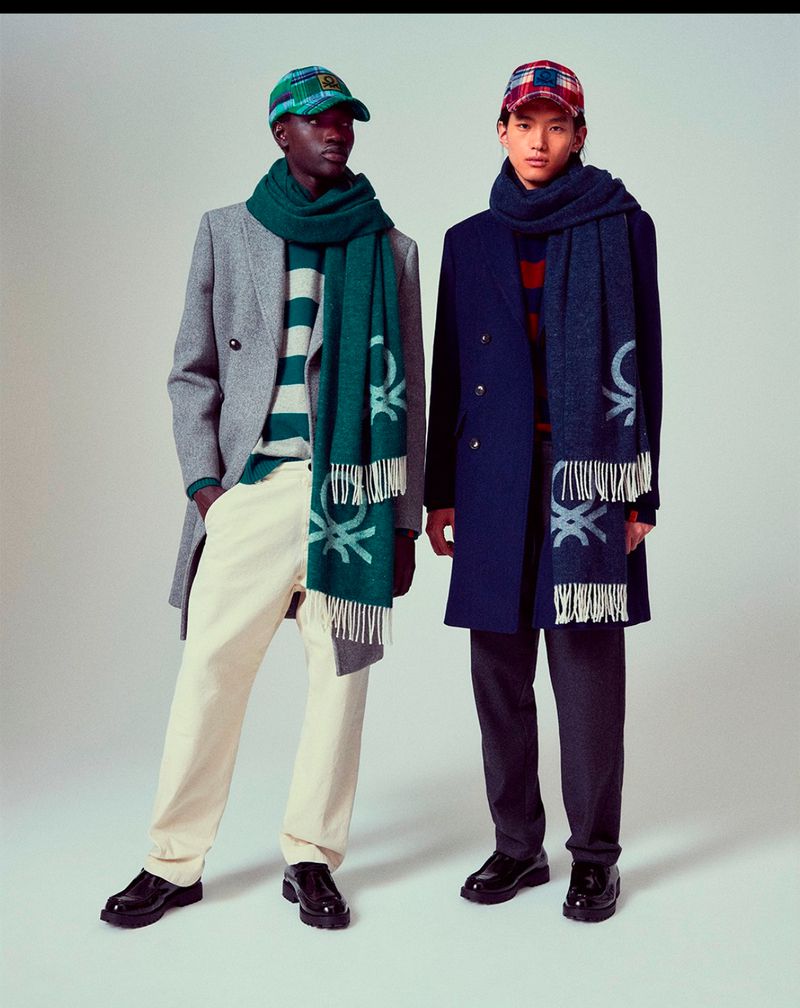 Two young adult male fashion models wearing coats, caps, and scarves. One wears white pants and the other gray pants in a studio setting.