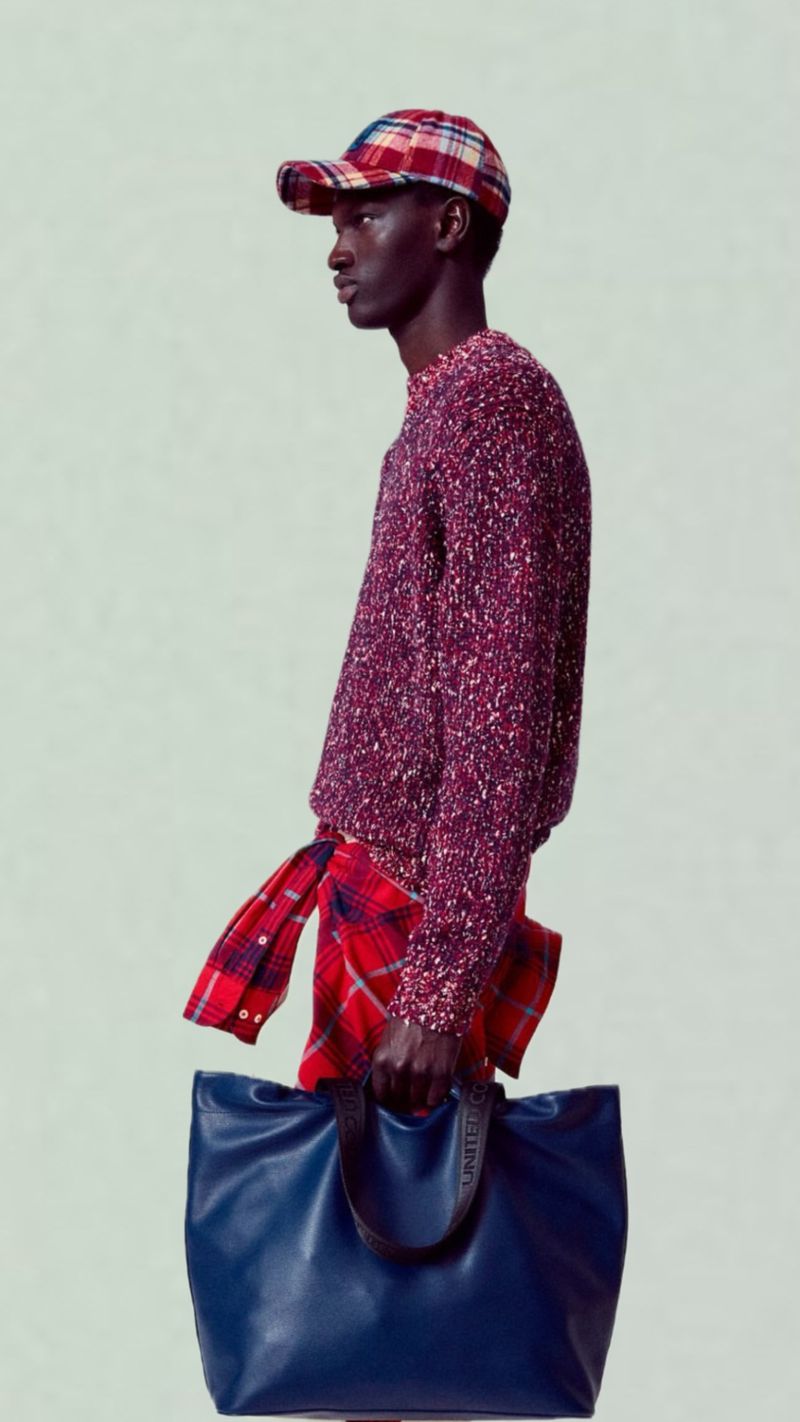 Profile of a male fashion model in a red sweater, flannel and ballcap carrying a navy tote bag in a bright studio setting.