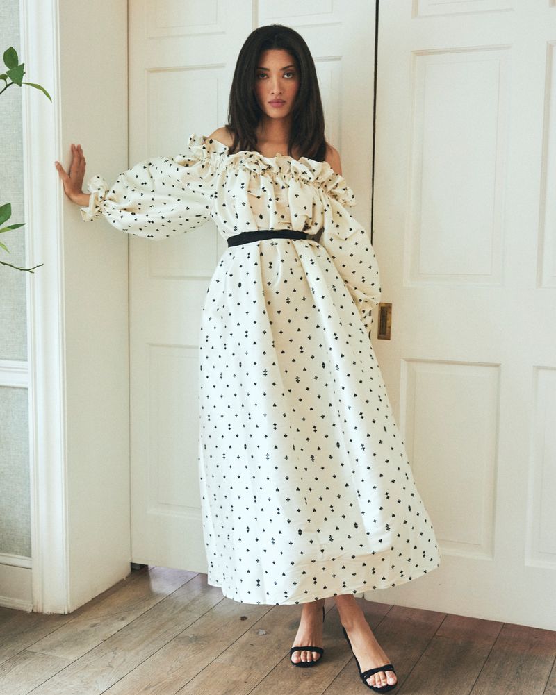 A full shot of a woman posing indoors in a white off-the-shoulder dress with black polka dots, a black belt, and black sandals.