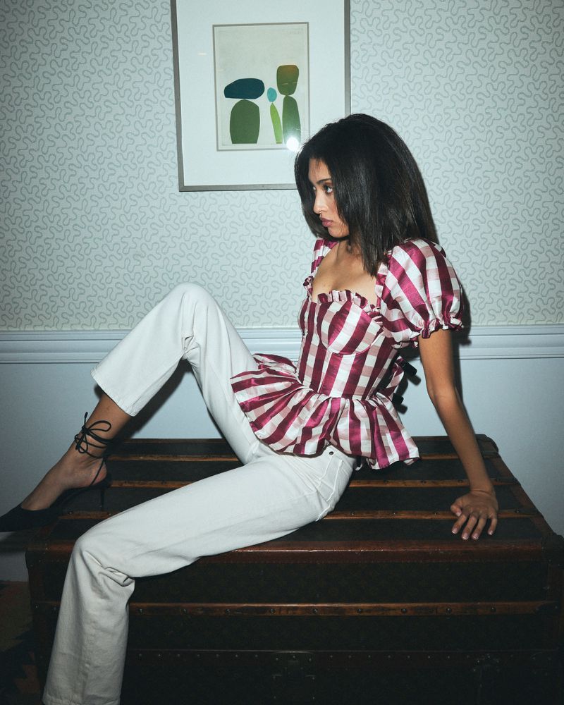 A woman in a red-and-white striped top and white jeans poses on a dark wood chest in an indoor setting with patterned wallpaper.