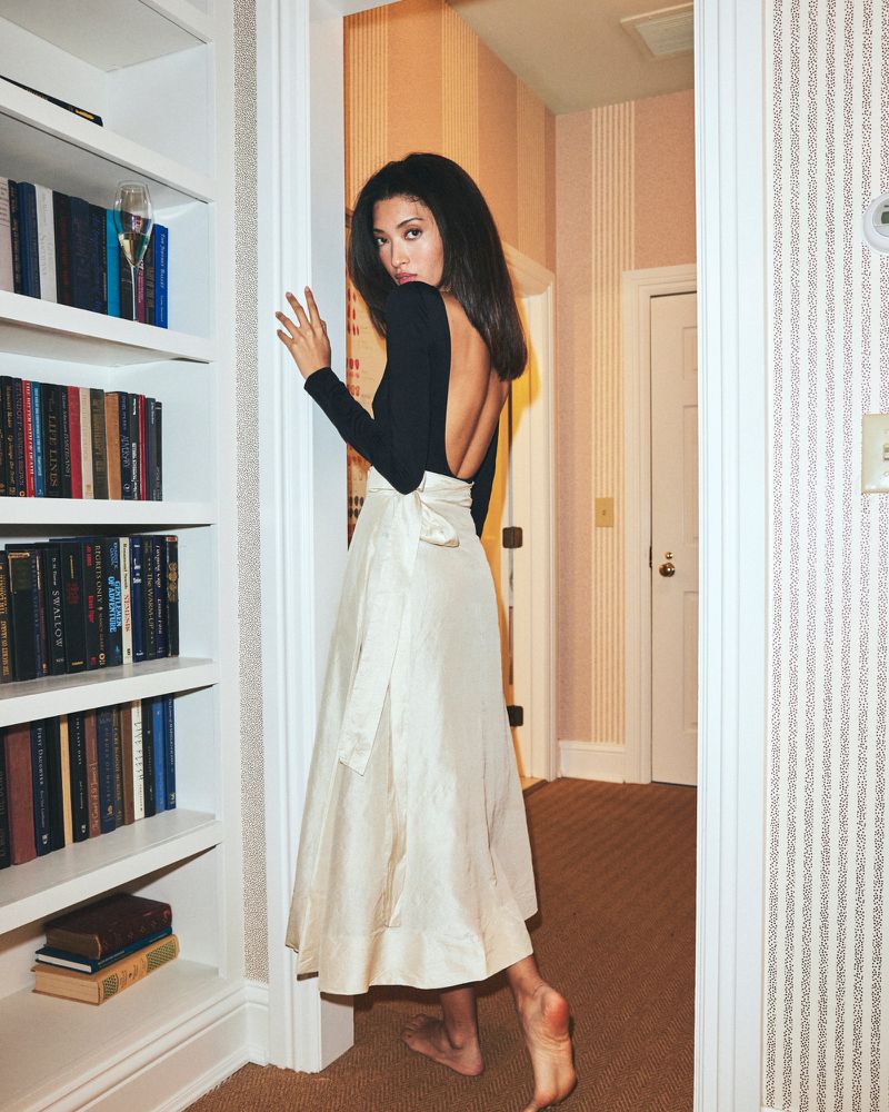 Elegant woman in a black top and ivory skirt stands beside a bookcase, creating a poised and stylish image, indoor setting with wallpaper.