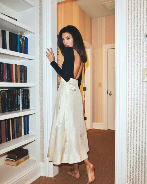 Elegant woman in a black top and ivory skirt stands beside a bookcase, creating a poised and stylish image, indoor setting with wallpaper.