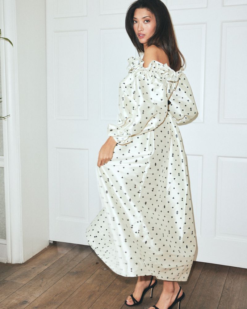 Model wearing an off-the-shoulder patterned white maxi dress posing against white panelled doors