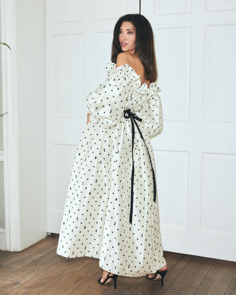 Elegant woman in off-shoulder dress with black ribbon detail poses indoors against a white paneled door and hardwood floor.